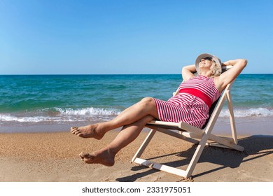 Smiling elderly woman is sitting in a sun lounger on a sandy beach near the sea on a sunny day. The lady enjoys nature and relaxation. Active lifestyle and health. Space for text. - Powered by Shutterstock