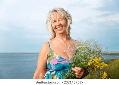 Smiling elderly woman in a summer sundress holding flowers in a field. Love for nature and relaxation.