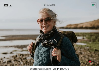 Smiling elderly woman with sunglasses and backpack by the coast. Outdoor adventure, coastal scenery, and joyful expression captured in a candid moment. Elderly woman backpacker vacation. - Powered by Shutterstock