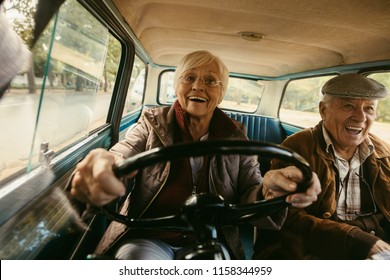 Smiling Elderly Woman In Warm Clothing Driving Car With Husband Sitting In Passenger Seat. Old Couple Having A Great Time On Their Drive.