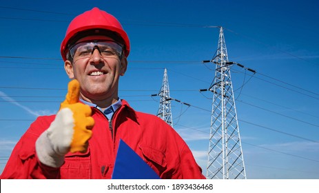 Smiling Electric Utility Worker Giving Thumbs Up Next To Electrical Transmission Towers. Portrait Of  Engineer In Red Hardhat And Coveralls. A Line Of High Voltage Electrical Transmission Towers.