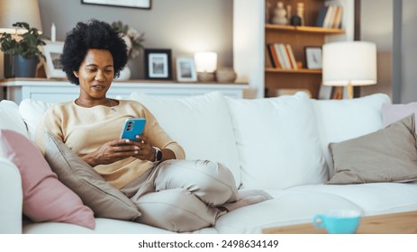 A smiling elegant African-American female using her smartphone while sitting on the cozy sofa in the living room. Beautiful mixed race woman browsing internet on cellphone in home living room. - Powered by Shutterstock