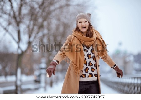 smiling elegant female in brown hat and scarf in camel coat with gloves outside in the city in winter.