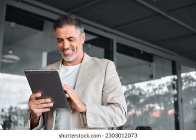 Smiling elegant mid aged business man wearing suit standing outside office holding digital tablet. Mature businessman professional using fintech device working on modern technology gadget. - Powered by Shutterstock