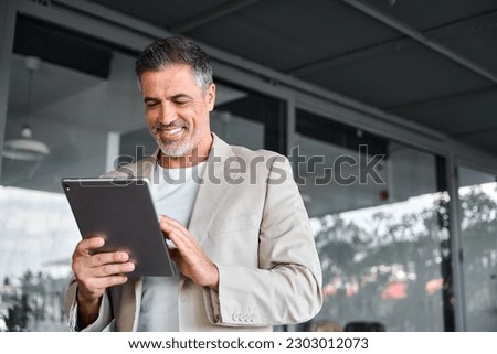 Smiling elegant mid aged business man wearing suit standing outside office holding digital tablet. Mature businessman professional using fintech device working on modern technology gadget.