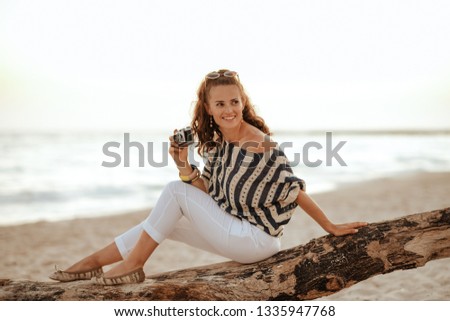 smiling elegant solo traveller woman in white pants and striped blouse with retro film photo camera looking aside while sitting on a wooden snag on the beach in the evening.
