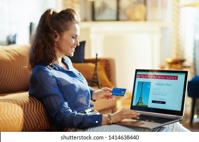 Smiling Elegant Woman With Opened On Laptop Online Travel & Booking Agency Site Holding Blue Credit Card In The Modern House In Sunny Summer Day. A Fake Website Is Made For Illustrative Purposes.