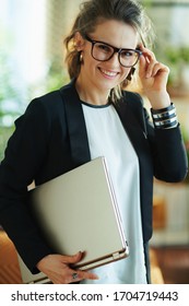 Lächelnde elegante Frau in weißer Bluse und schwarze Jacke in Gläsern mit geschlossenem Laptop im modernen Haus an sonnigen Tag.