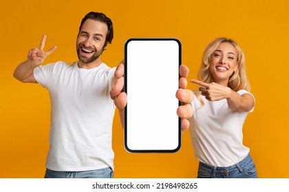 Smiling Emotional Millennial European Man And Lady Show Finger To Big Phone With Blank Screen, Peace Sign, Isolated On Yellow Background, Studio. Fun Together, Couple Enjoy New App, Great Ad And Offer