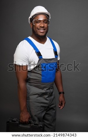 Smiling engineer in protective helmet and transparent glasses stands against gray wall holding suitcase with tools in hand. Satisfied black engineer with box professional tools in his hand looks ahead