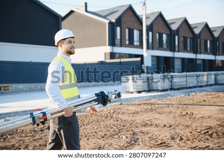 Smiling engineer wearing a hardhat and high visibility vest, carrying surveying tools while walking on a new residential construction site