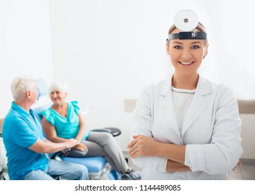 Smiling ENT Doctor With Her Senior Patients In The Background, Focus On Foreground At Medical Room