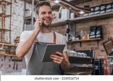 Smiling entrepreneur in apron near counter of his cafe using digital tablet and talking on smartphone - Powered by Shutterstock