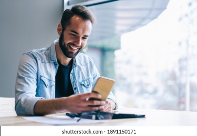 Smiling entrepreneur with beard in casual clothes sitting at table with tablet and browsing smartphone in modern workspace with large window in daytime - Powered by Shutterstock