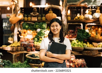 Smiling entrepreneur holding a digital tablet standing at an outdoor market. Asian woman in an apron looking at camera while standing against a street food market. - Powered by Shutterstock