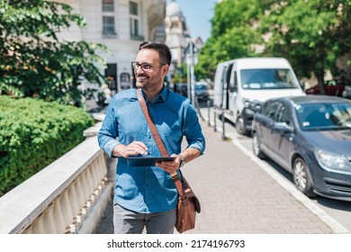 Smiling Entrepreneur Using Digital Tablet. Male Executive Commuter Is Looking Away While Walking On Sidewalk. He Is Wearing Formals In The City During Sunny Day.