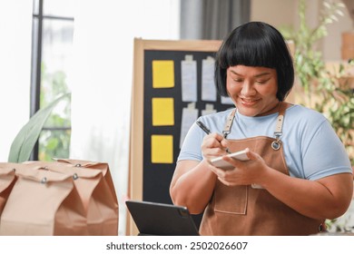 smiling entrepreneur wearing apron writes notes preparing food delivery orders in modern, well-lit workspace. includes tablet, packaging bags indicating small business or home-based food delivery - Powered by Shutterstock