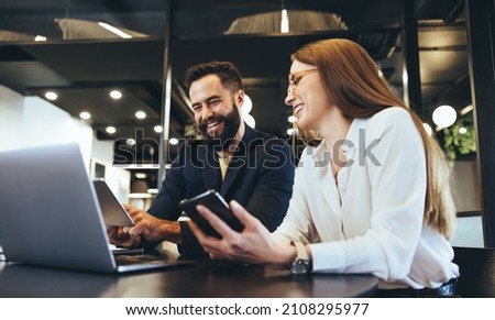 Smiling entrepreneurs using wireless technology while working together in an office. Two happy businesspeople looking at a digital tablet while sitting at a table in a modern workspace.
