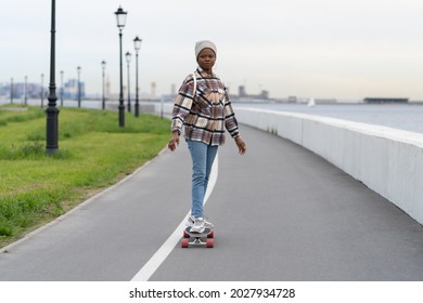 Smiling ethnic black woman on skateboard. Happy skateboarder female enjoy longboarding in early spring. Cheerful african american girl skating city road, urban deck space with sea or river view alone - Powered by Shutterstock