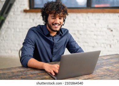 Smiling Ethnic Indian Man Wearing Wireless Headset Using Laptop For Virtual Connection, Talking With Colleagues, Indian Male Student Involved Webinar. Latin Employee Of Customer Service Support