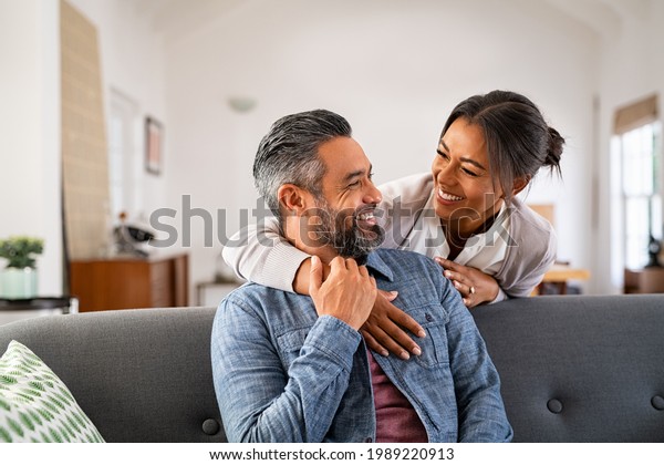 Smiling ethnic woman hugging her husband on the
couch from behind in the living room. Middle eastern man having fun
with his beautiful young wife on the couch. Mid adult indian man
with latin woman.
