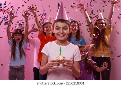 Smiling european boy holding birthday cupcake with candle with background of celebrating friends in flying confetti. Boys and girls wearing party hats. Party and holiday. Pink background. Studio shoot - Powered by Shutterstock