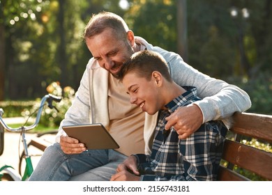 Smiling european father and teenage son with cerebral palsy watch digital tablet and rest on bench in park. Family spending time together. Disability care, treatment and rehabilitation. Warm sunny day