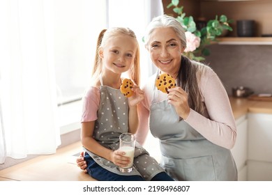 Smiling European Granddaughter And Old Grandma In Aprons Enjoy Cookies With Chocolate With Glass Of Milk At Kitchen Interior. Homemade Sweets, Cooking Together, Baking For Family At Home And Breakfast