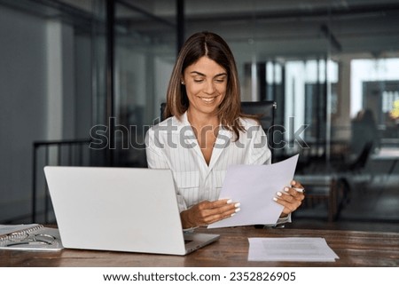 Smiling European Latin business woman accountant analyst holding documents, work at laptop computer doing online trade market tech research. Focused Hispanic businesswoman with paperwork in office.