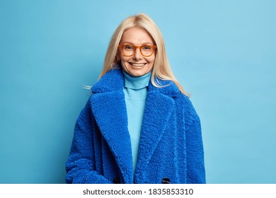 Smiling European woman with blonde hair dressed in blue winter coat has happy mood wears eyewear poses in studio. Fashionable middle aged blonde lady in outerwear going to have walk with family