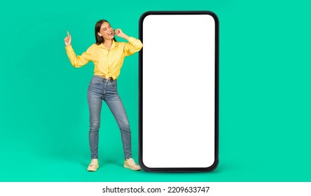 Smiling european young woman in headphones have fun, sing, enjoy music near big smartphone with blank screen, isolated on green background, studio. Ad and offer, website and new app for relaxation - Shutterstock ID 2209633747