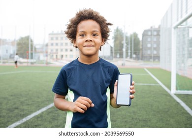 Smiling And Excited Boy Showing Smartphone Empty Screen. Looking To Camera. Cellphone Display Mockup Mobile App Advertisement. African American Kid In Football Field. Blank Screen
