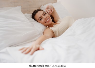 Smiling excited fun young woman in casual casual clothes lying in bed reach hand to camera you rest relax spend time in bedroom lounge home in own room house wake up dream be lost in reverie good day