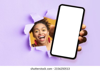 Smiling Excited Pretty Young Black Woman With Open Mouth Looks Through Hole In Purple Paper And Shows Smartphone With Blank Screen, Studio. Huge Sale, Online Win, Great Offer, App And New Technology