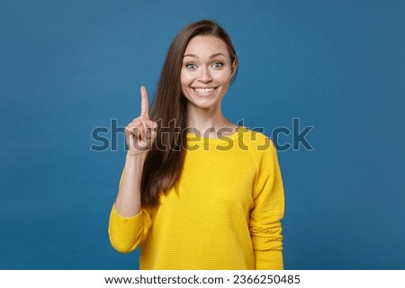 Smiling excited young brunette woman 20s wearing yellow casual clothes posing standing holding index finger up with great new idea looking camera isolated on blue color background, studio portrait
