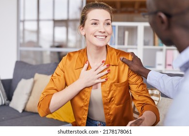 Smiling excited young woman in orange blouse sitting on sofa and thanking therapist while black therapist touching her shoulder and supporting her at psychology session - Powered by Shutterstock