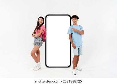 Smiling exited high school pupils students teenagers classmates boy girl friends in casual with bags pointing at big mock up phone with blank display area isolated on white background empty space.