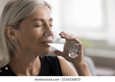 Smiling face of elderly lady relieving stress having glass of natural cold filtered water in dry hot summer day. Mature woman has healthy habit for easy skin body care hydrating drink enjoy every sip