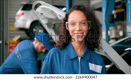 Smiling face of Latin woman car mechanic in uniform standing looking at camera at autocar
