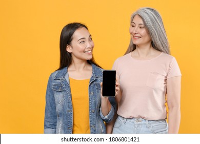 Smiling Family Asian Women Mother Daughter In Casual Clothes Posing Hold Mobile Phone With Blank Empty Screen Mock Up Copy Space Looking At Each Other Isolated On Yellow Background Studio Portrait