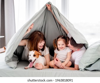 Smiling Family With Dog Under The Blanket In The Bed. Beautiful Morning Moments Of Mother, Father And Little Daughter With Pet Under Coverlet. Young Mom And Dad Looking At Their Kid