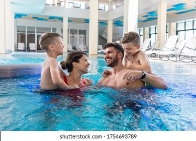 Smiling Family Of Four Having Fun And Relaxing In Indoor Swimming Pool At Hotel Resort.