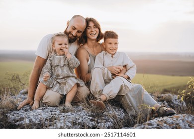 Smiling family of four sitting together on rocky ground, enjoying a sunset in a rural landscape. Capturing moments of joy, laughter, and togetherness in a serene outdoor setting. - Powered by Shutterstock