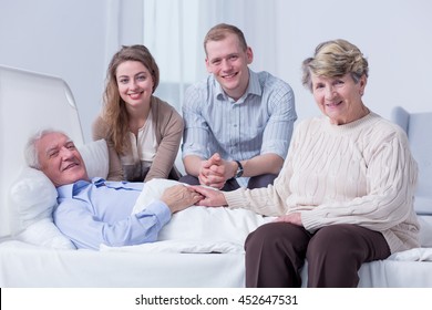 Smiling Family Members Surrounding The Bed Of An Old Man In A Very Bright Hospital Room