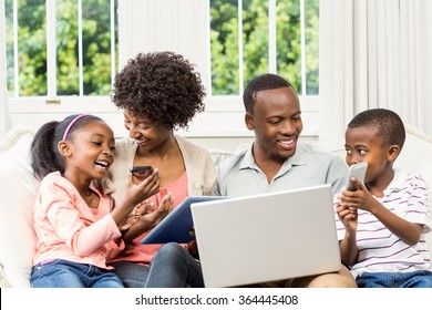 Smiling Family Sitting On The Couch Together Using Laptop And Tablet And Smartphone