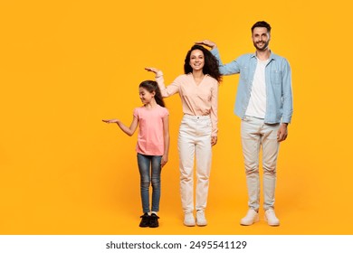 A smiling family stands against a bright yellow background, measuring their heights by holding their hands out. The daughter stands to the left, her mother in the middle, and the father on the right. - Powered by Shutterstock