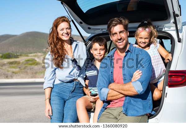Smiling family with two kids sitting in car trunk\
and looking at camera. Happy children enjoying with mother and\
father a road trip while sitting in car back. Portrait of cheerful\
young family in auto