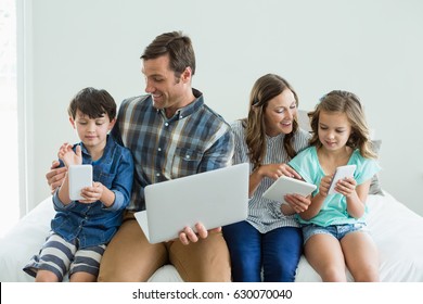 Smiling Family Using Laptop, Digital Tablet And Mobile Phone In Bedroom At Home