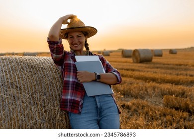 Smiling farmer woman with tablet, folders, and straw hat posing in her hay field amidst rolled bales. Sunsets and fields provide a picturesque backdrop - Powered by Shutterstock