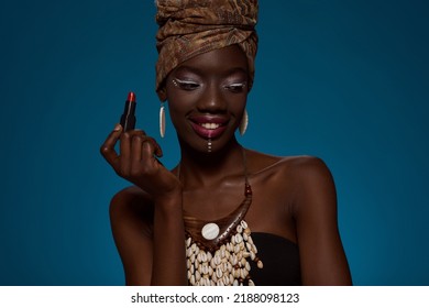 Smiling Fashionable Black Girl With Lipstick. Pretty Young Slim Woman Wearing Traditional African Outfit And Accessories. Female Beauty. Isolated On Blue Background. Studio Shoot. Copy Space